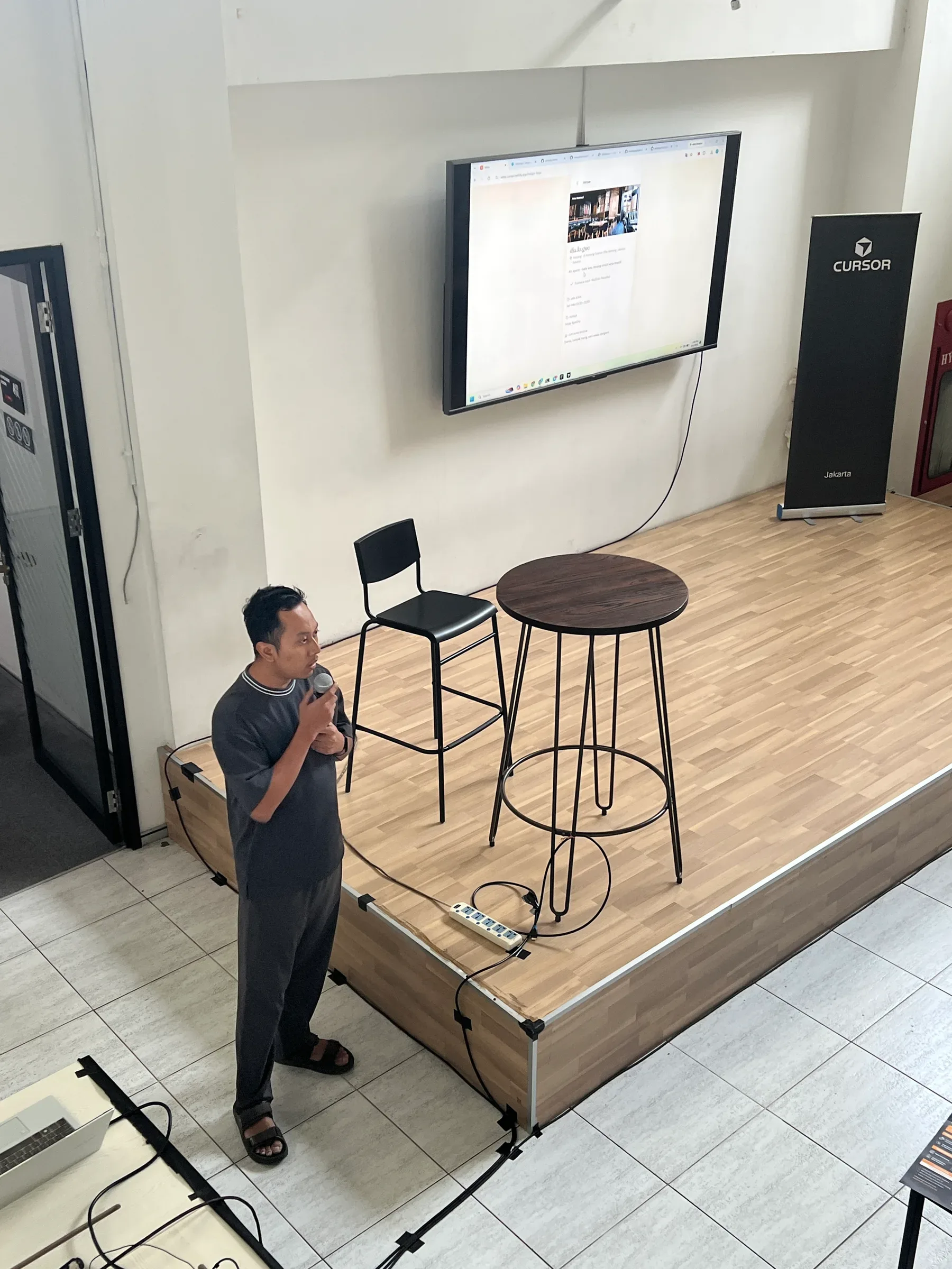 Speaker holding a microphone beside a presentation screen at the Cursor Jakarta workshop