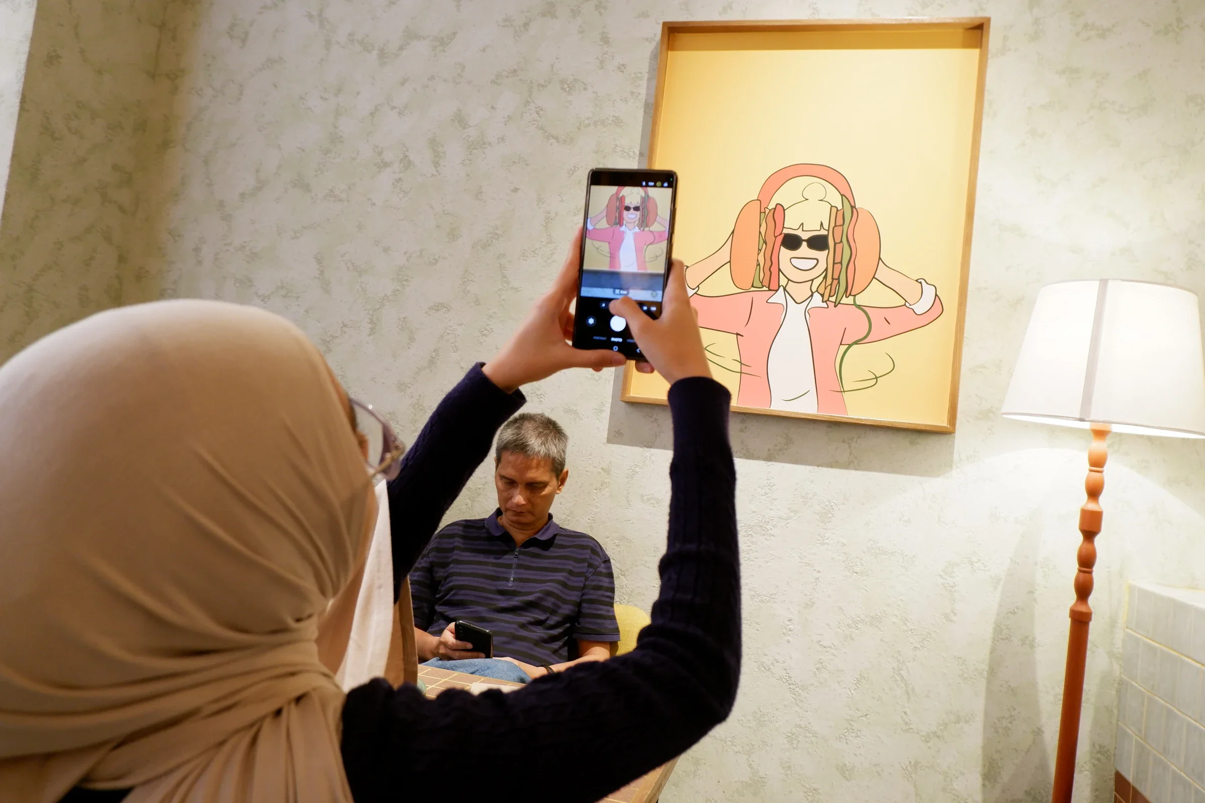 A person raising a phone toward colorful wall art, with another person seated below